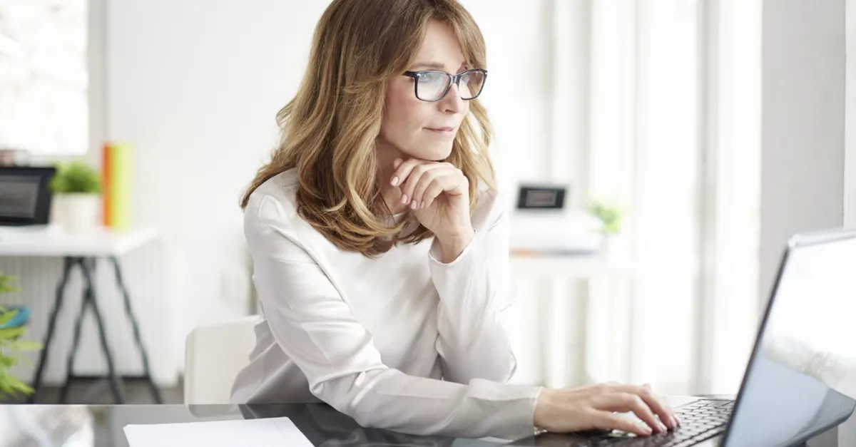 Individual seated at a glass desk using a laptop in a bright modern office with large windows, papers, and office supplies visible.