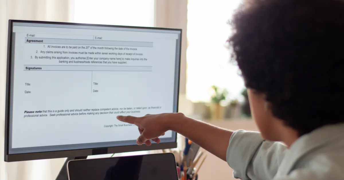 Individual seated at a desk pointing at a computer monitor displaying an agreement document with sections for email, signatures, and terms, with a laptop and papers