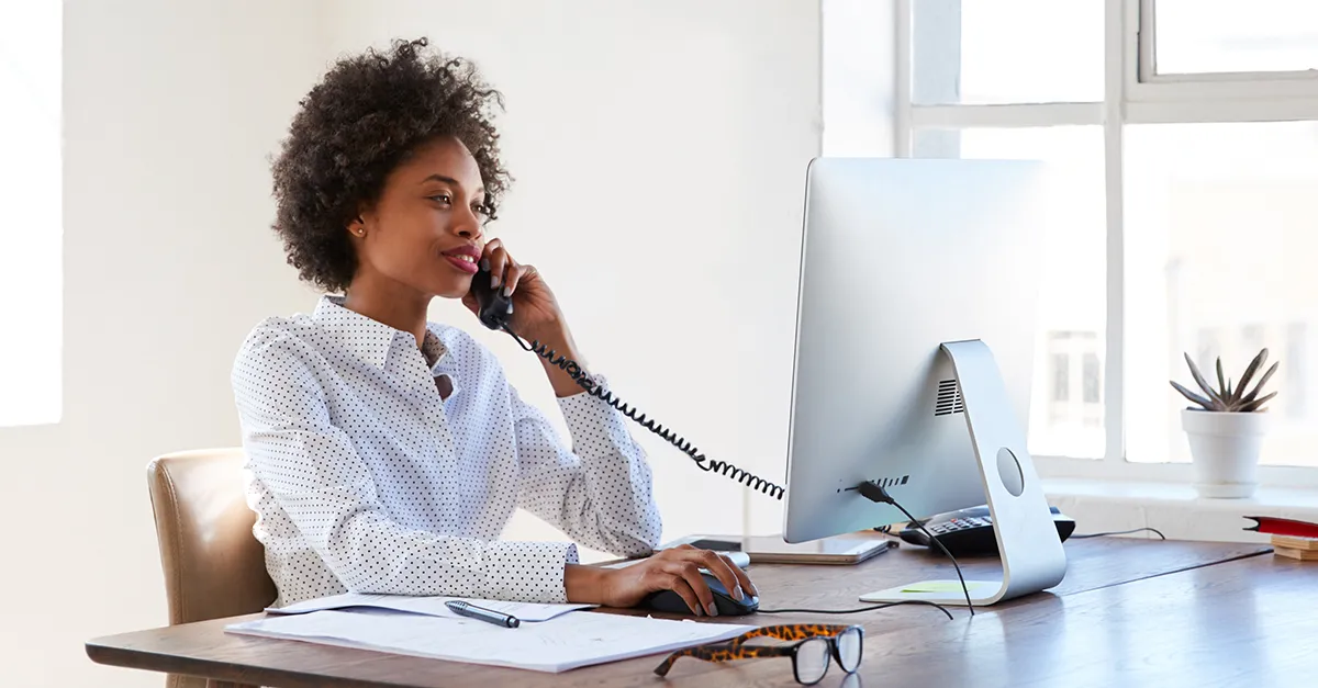 Individual seated at a wooden desk in a bright office, speaking on a corded phone while using a desktop computer, with papers, pen, and eyeglasses placed on the desk.