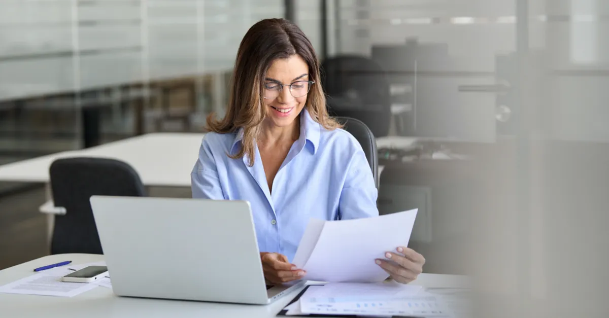 Individual seated at a desk in a modern office, holding printed documents while working on a laptop, with additional papers and a pen placed on the table.