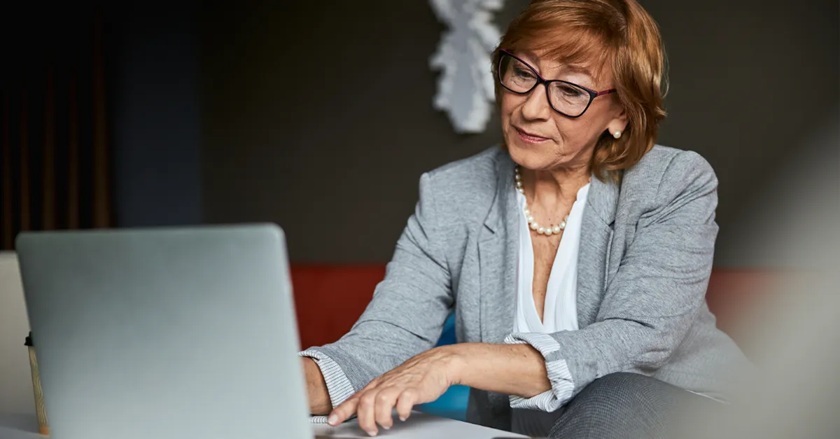 Person in a business suit working on a laptop at a desk with a smartphone nearby.