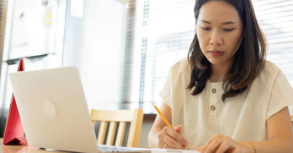 Person writing in a notebook while working on a laptop at a home workspace.