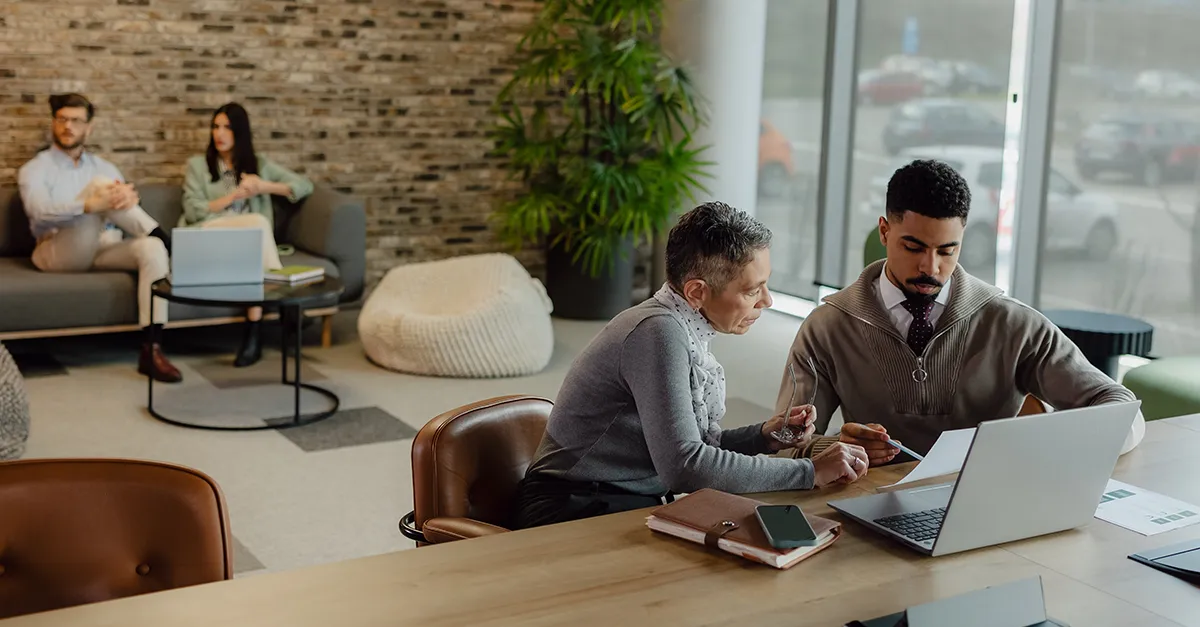 People working together in a modern office lounge, with two individuals discussing documents at a table and others seated in the background.