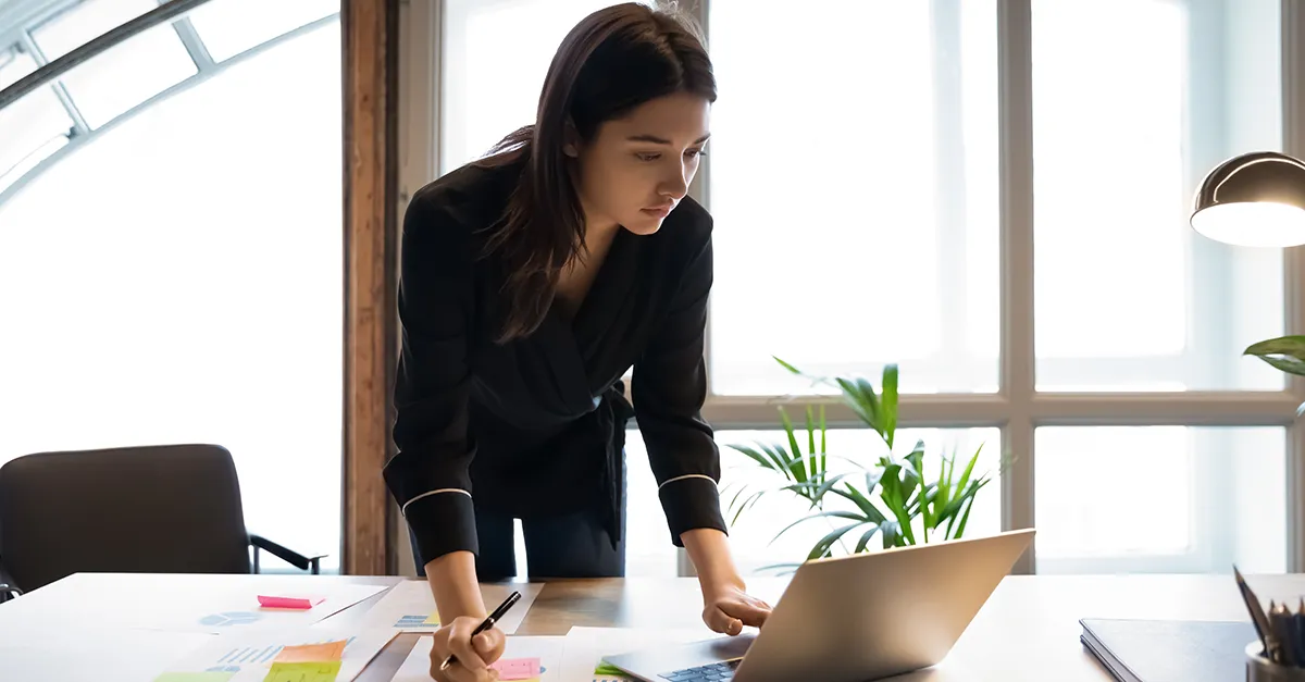 Person reviewing documents and working on a laptop while standing at a desk in a modern office.
