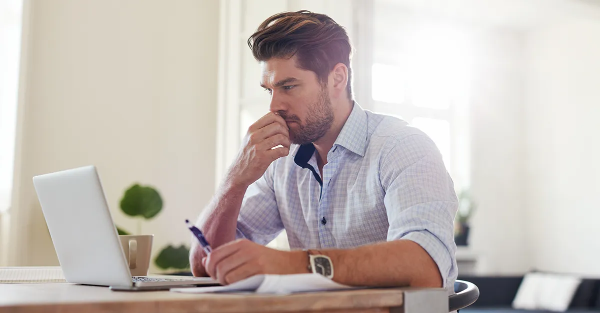 Person writing in a notebook while working on a laptop at a bright home workspace