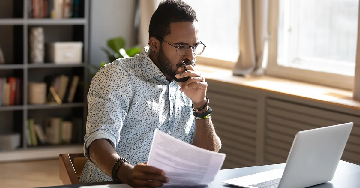Person reviewing a printed document while working on a laptop in a home office.