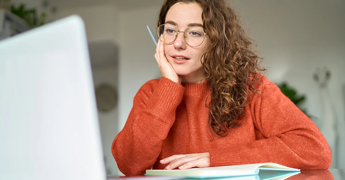 Person sitting at a desk, holding a pen and looking at a laptop while writing in a notebook.