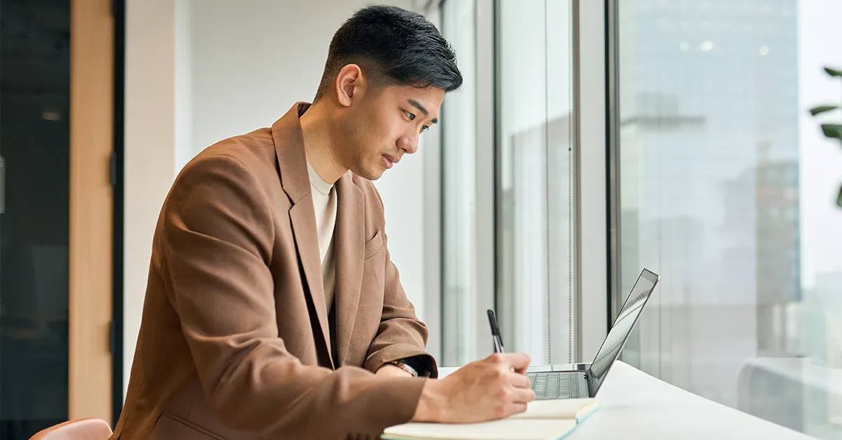 Person working at a desk, taking notes while using a laptop near a large window overlooking city buildings.