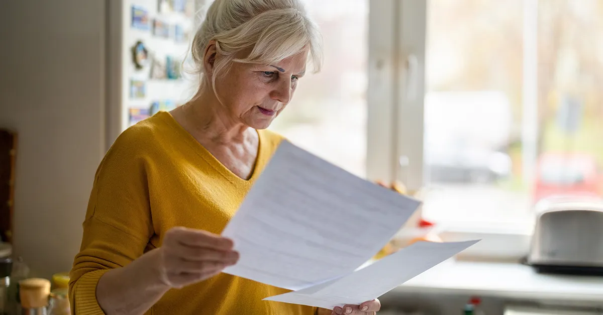 An older woman standing in her kitchen reviewing documents in her hands.