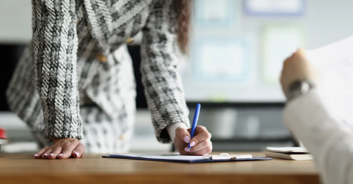 A businesswoman leaning over a desk while signing documents during a meeting in an office setting.