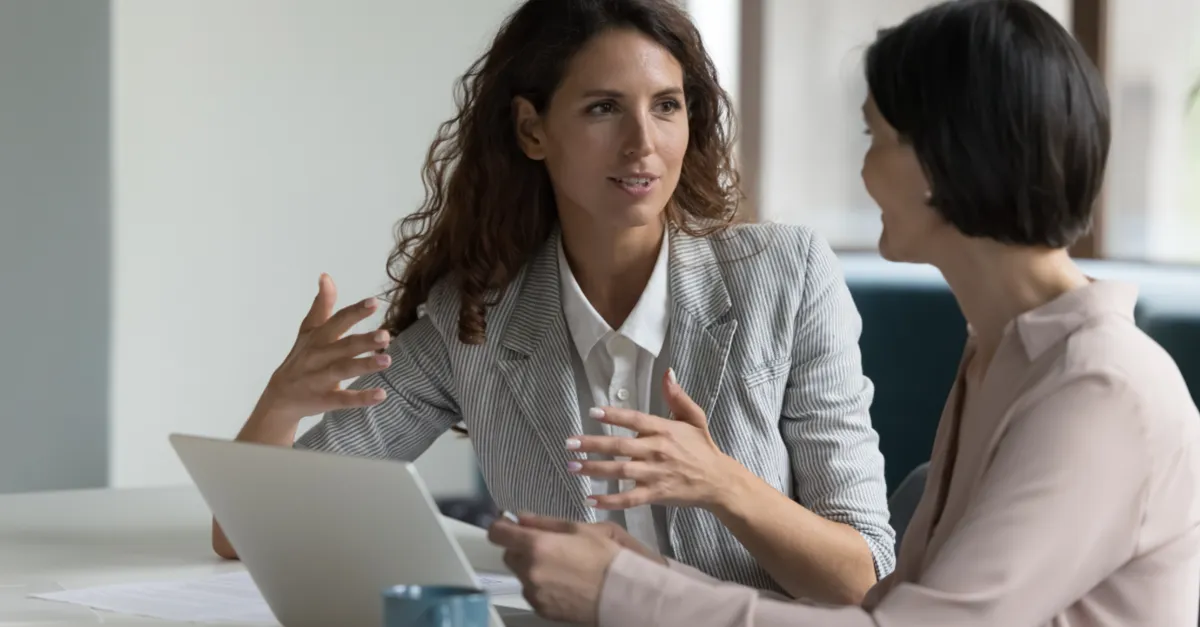 Two colleagues discussing work together at a desk with a laptop during an office meeting.