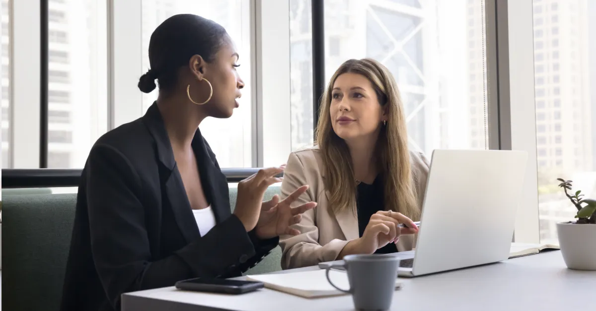 Two colleagues sitting at a table in a modern office discussing work, with a laptop and notebooks in front of them.