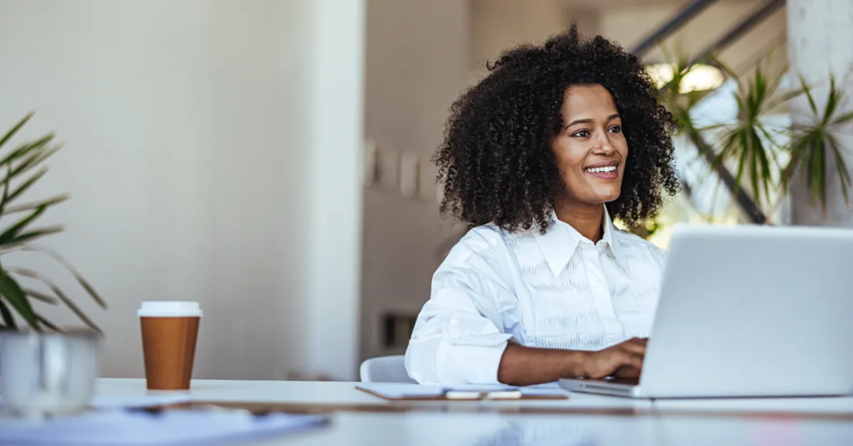 A person working on a laptop at a desk in a modern office with documents and a coffee cup nearby.