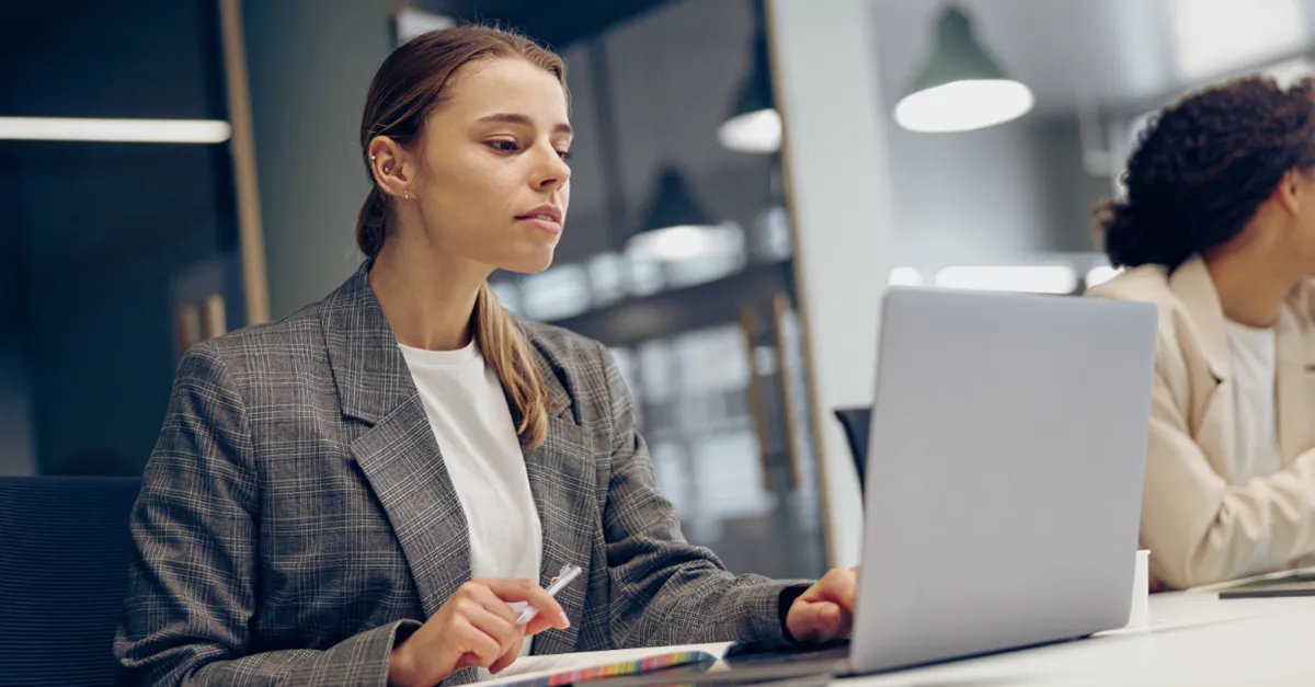 A person sitting at a desk in an office, using a laptop and holding a pen with documents in front of them.