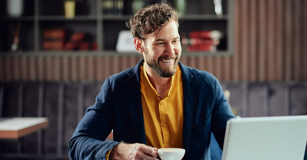 Man working on a laptop in a cafe while holding a cup of coffee.
