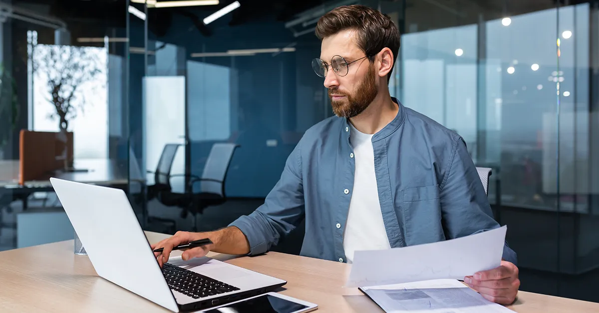 rofessional working at a desk with a laptop and holding documents in a modern office environment