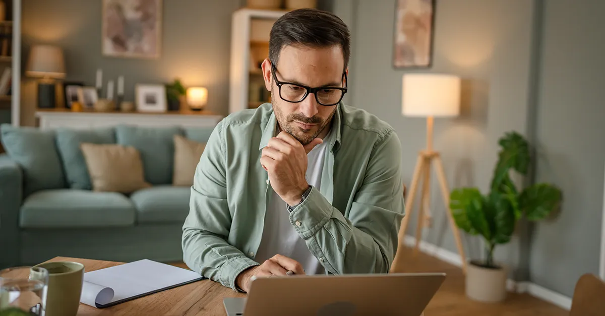 Person working on a laptop at a desk in a home office, with documents and a coffee nearby.