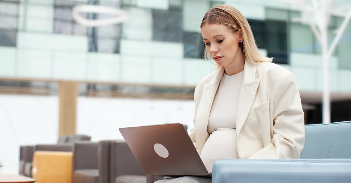 Pregnant professional working on a laptop while seated in a modern office or lounge setting.