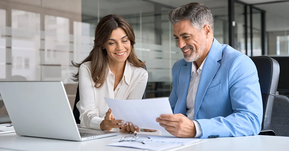 Two professionals seated at a desk reviewing documents together next to a laptop in a modern office.
