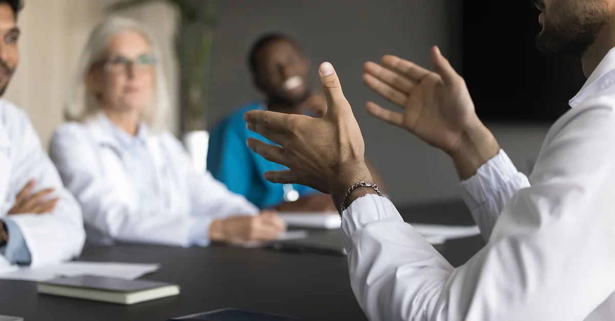 Medical professionals sitting around a table in a meeting while one person speaks and gestures during the discussion.