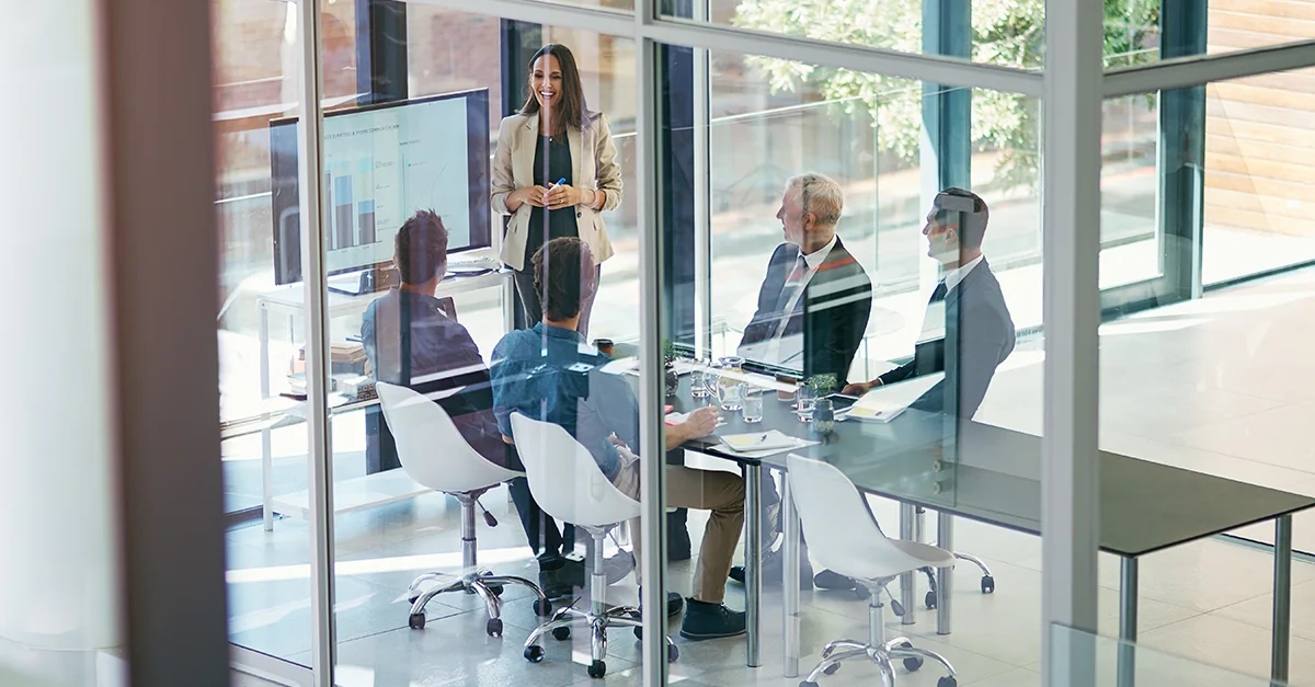 A team seated around a conference table in a glass meeting room while a presenter stands near a screen displaying charts.