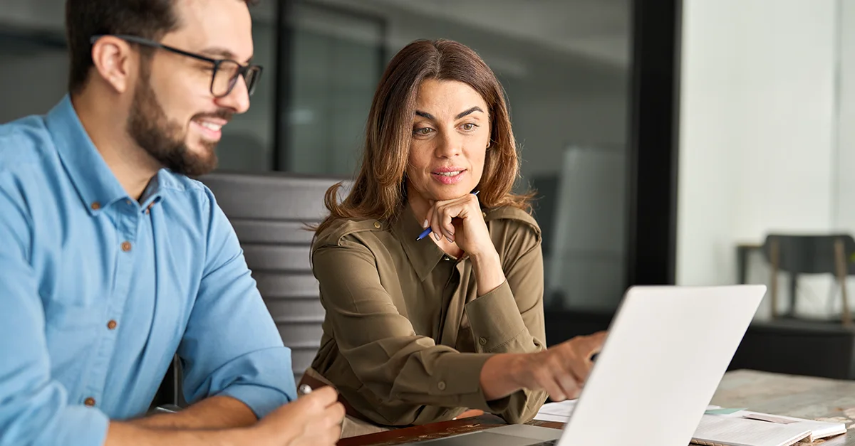 Two colleagues sitting together in an office, looking at a laptop while discussing information on the screen.