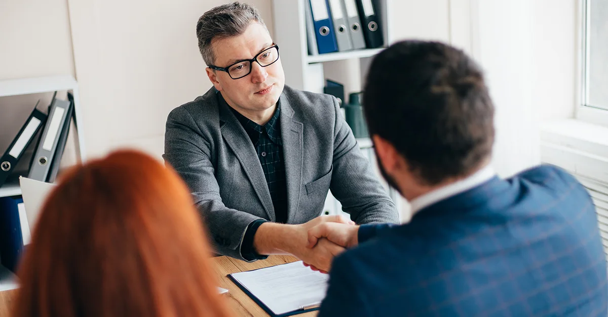 Two people seated across a desk shaking hands during a job interview or business meeting in an office.
