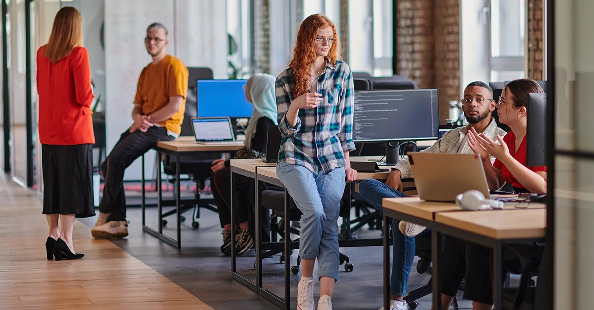 People working and talking together in a modern open‑plan office, with some seated at desks using laptops and others standing and having conversations.