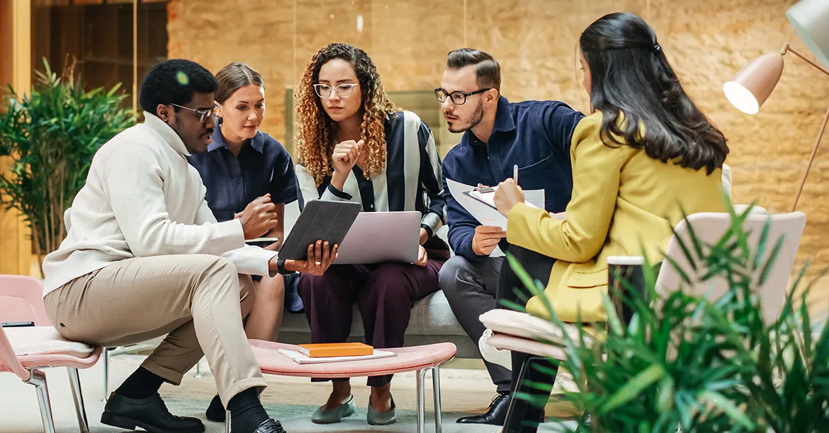 A group of colleagues sitting together in a modern office lounge area, reviewing documents and discussing work around a small table.