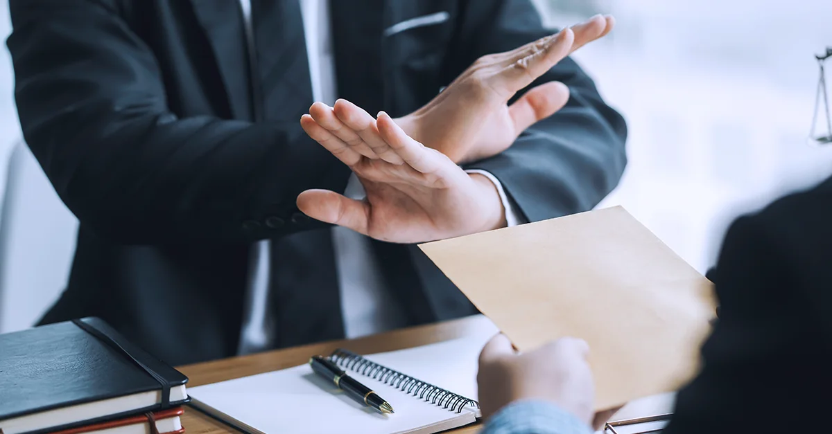 A person in business attire holding up both hands in a stopping gesture while another person offers an envelope across a desk.
