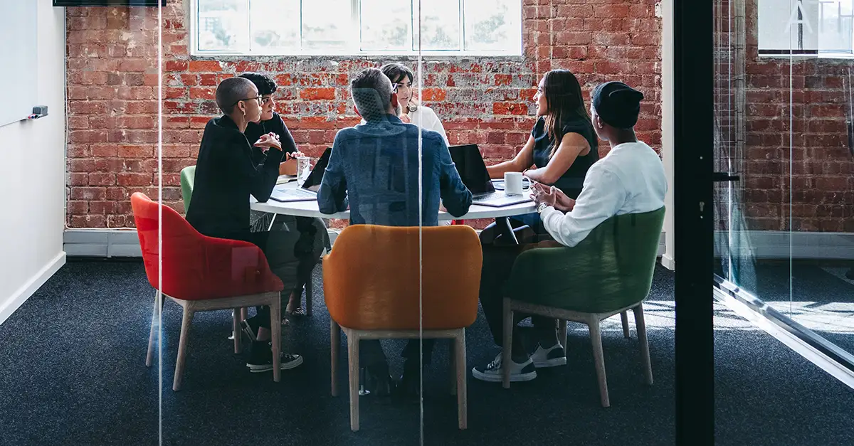 A group of colleagues sitting around a conference table in a modern glass‑walled meeting room, engaged in a discussion.
