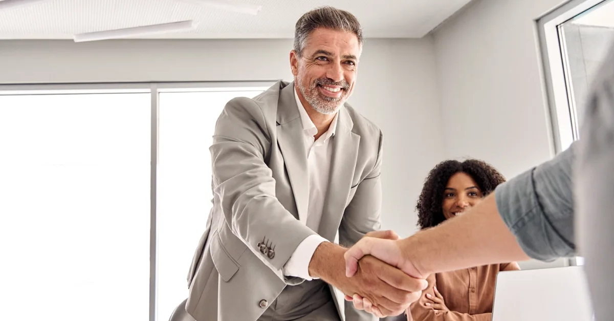 Two people shaking hands across a desk during an office meeting while another person sits nearby.