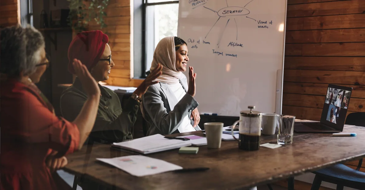 A group of people sitting around a table in an office, participating in a hybrid meeting with a video call on a laptop and strategy notes written on a whiteboard.