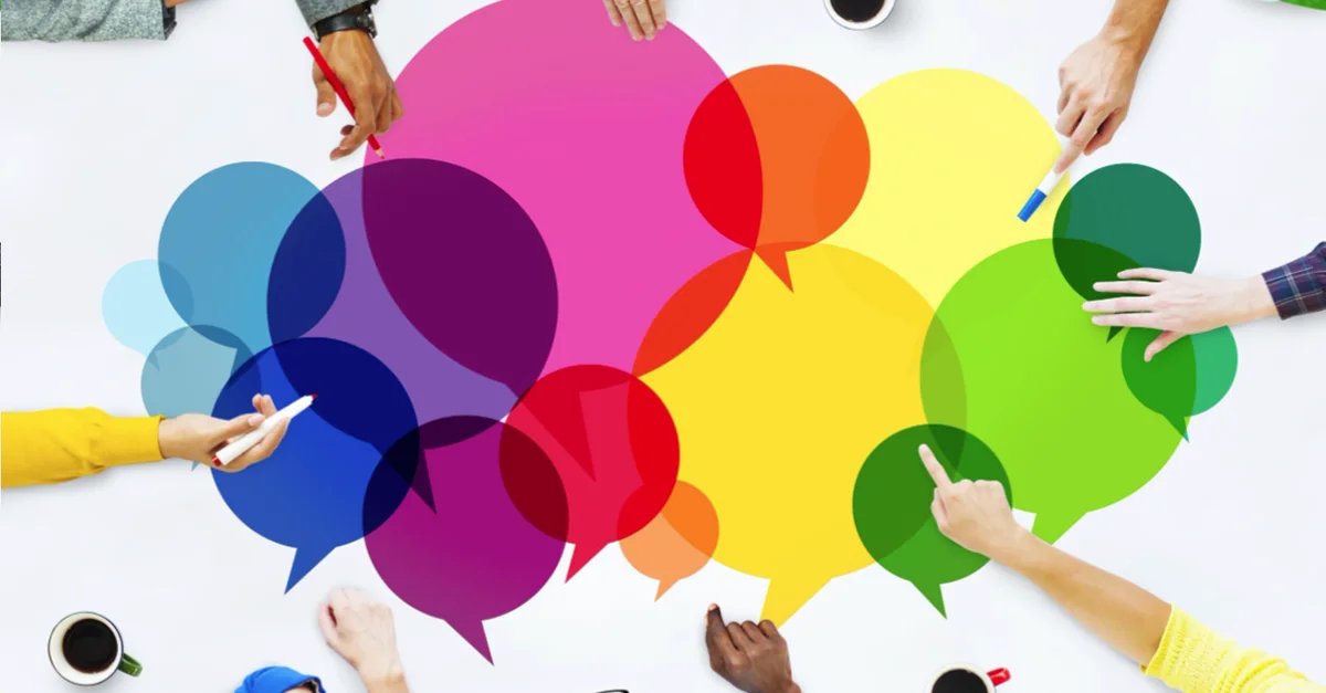A group of people gathered around a table with coffee cups and stationery, collaborating around a large display of colorful overlapping speech bubbles.