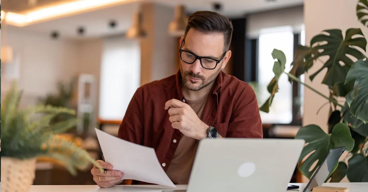 Individual sitting at a desk in a modern workspace, holding documents while working on a laptop with plants and office items in the background.