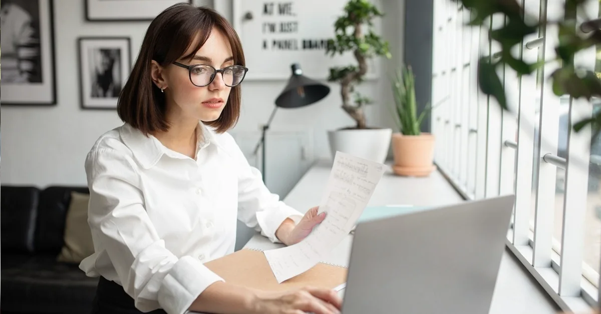 Individual standing at a desk by a large window, using a laptop and holding printed documents in a bright workspace with plants and framed artwork in the background.