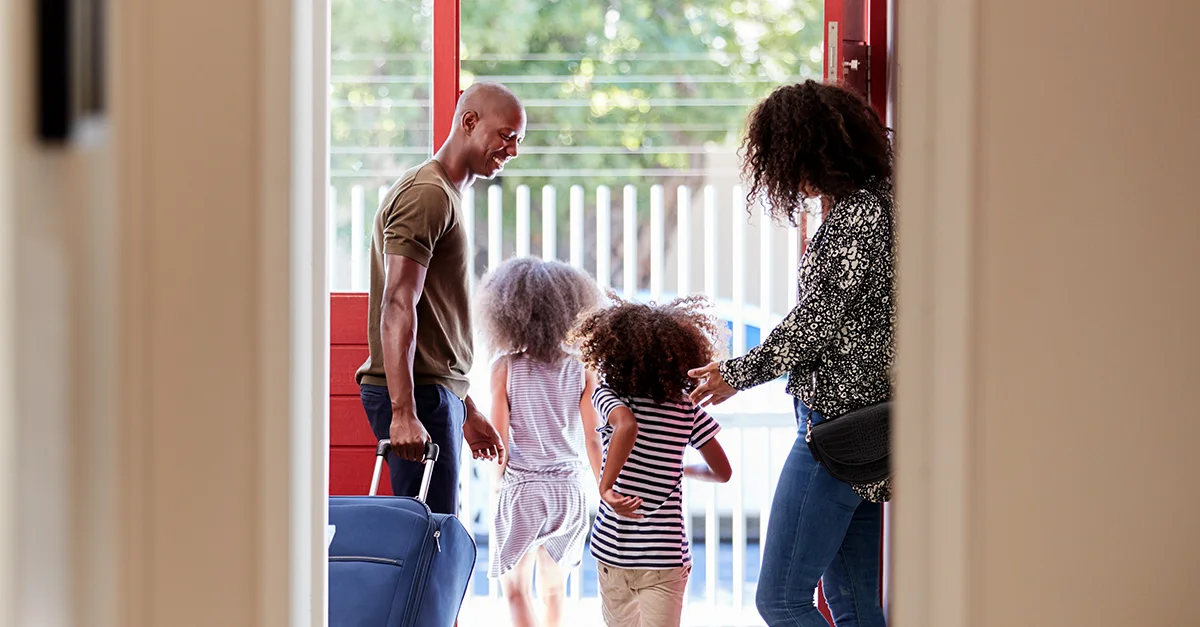 A family standing at the front door with luggage as they prepare to leave the house, with two children walking ahead.