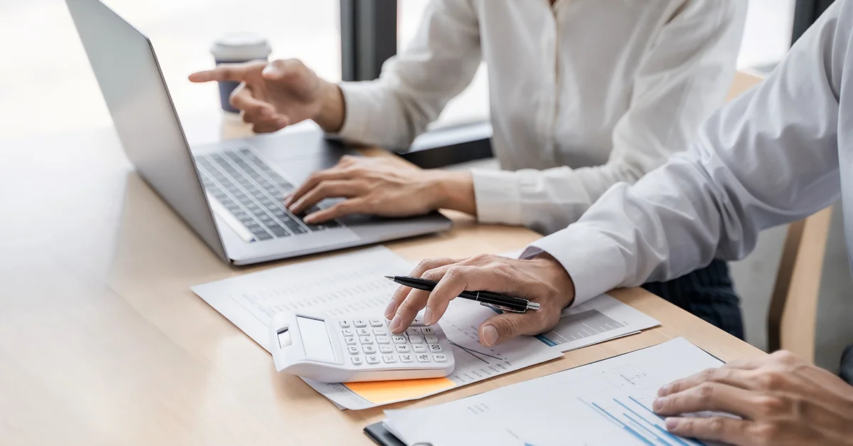 Two people sitting at a desk working together, using a laptop, calculator, and printed financial documents while discussing data.