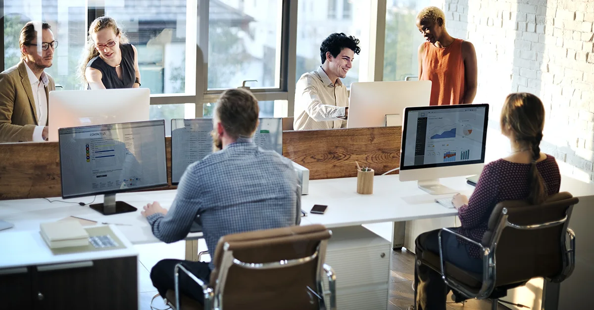 A group of people working at computers in a modern open‑plan office, with individuals collaborating and discussing tasks around shared desks.