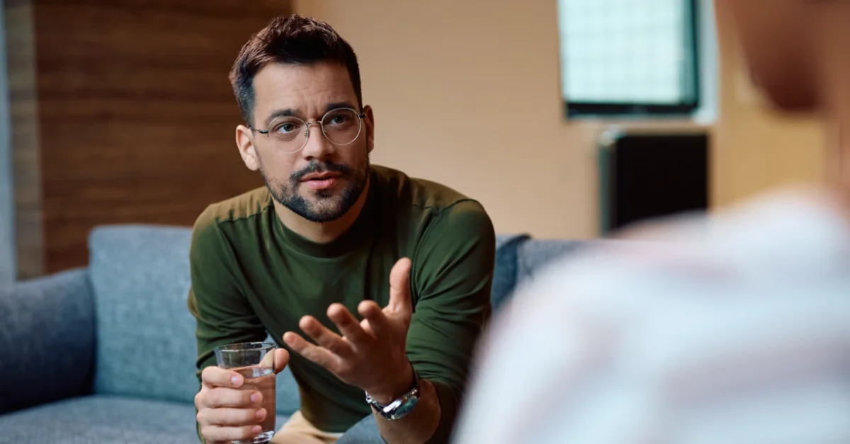 A person sitting on a couch, holding a glass of water, and gesturing with one hand while speaking to another individual who is taking notes in the foreground.
