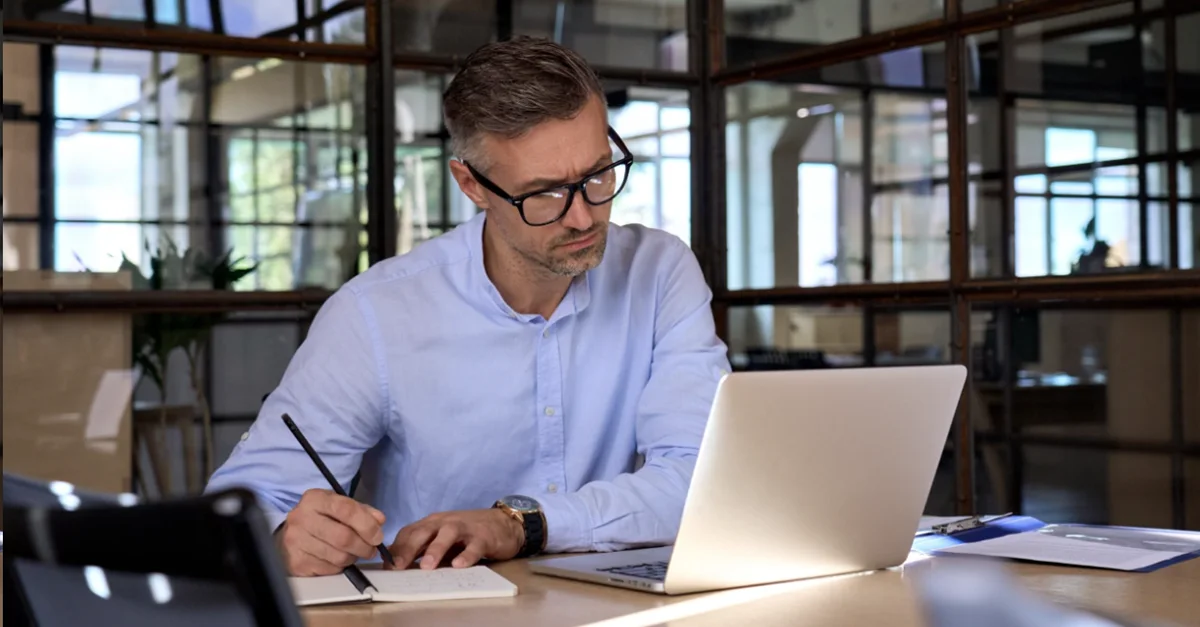 Individual sitting at a desk in a glass‑walled office, writing notes in a notebook while using a laptop in a well‑lit workspace.
