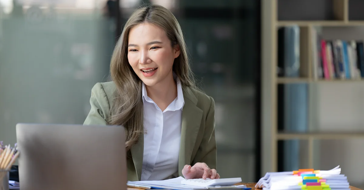Individual seated at a desk in an office environment, working with a laptop and reviewing documents alongside organized stacks of paperwork.