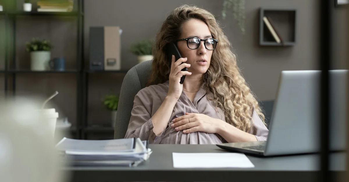 Individual sitting at an office desk, holding a phone to the ear with one hand and resting the other hand on the abdomen, with a laptop and documents on the desk.