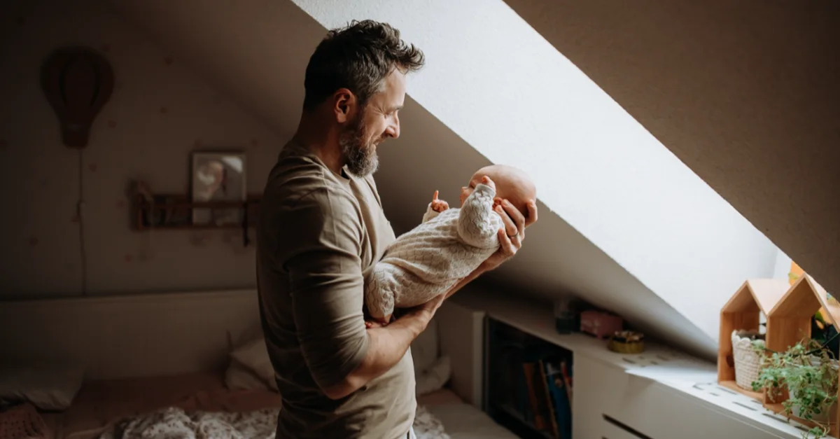 A person standing in a softly lit bedroom, holding a newborn baby near a slanted window with warm natural light filling the room.