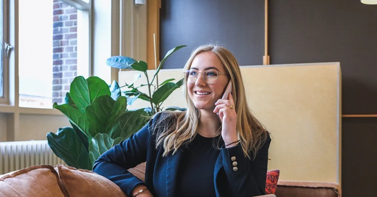 Individual seated on a couch in a modern office or lounge area, holding a hand near the chin with large indoor plants and soft natural light in the background.