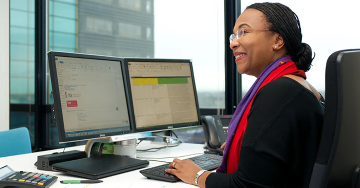 Individual seated at a desk in a modern office, working on two computer monitors displaying spreadsheets and emails, with a keyboard, mouse, and office supplies on the desk.