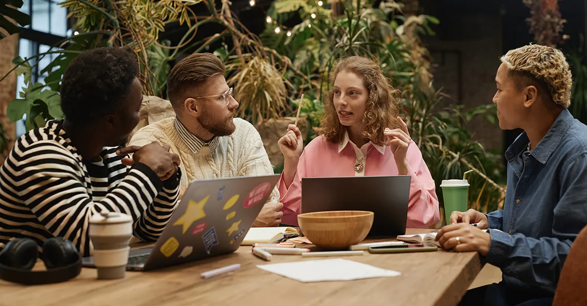 A group of individuals sitting around a wooden table in a workspace filled with plants, engaged in an active discussion with laptops, notebooks, and art supplies on the table.