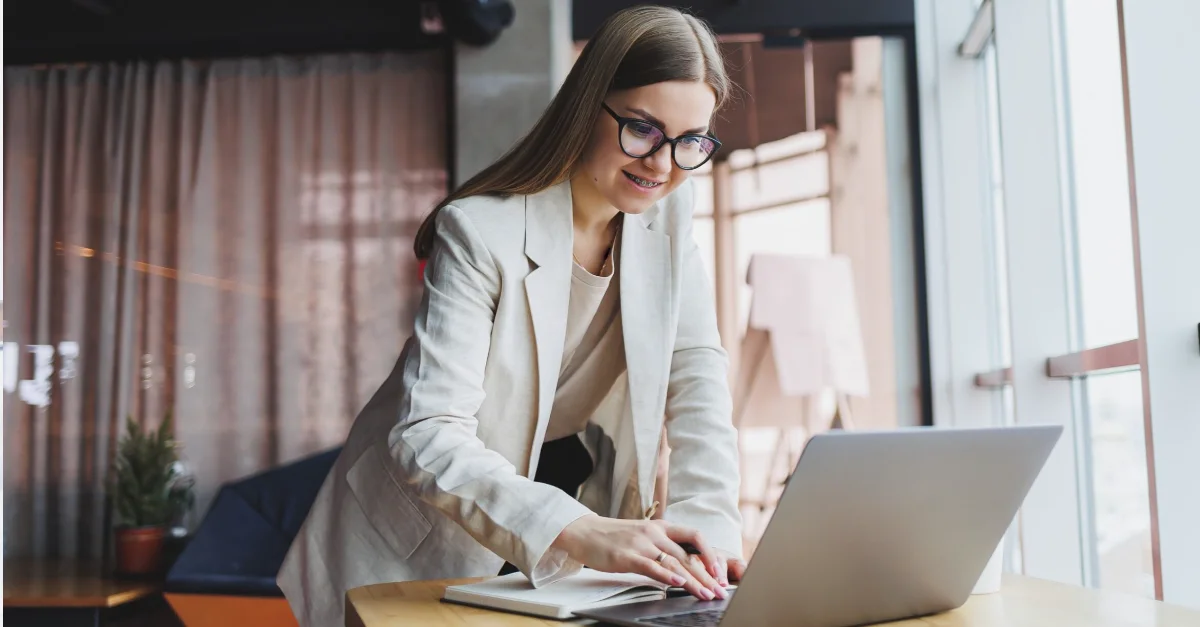 Individual standing at a high table and typing on a laptop in a modern indoor space, with large windows providing natural light.