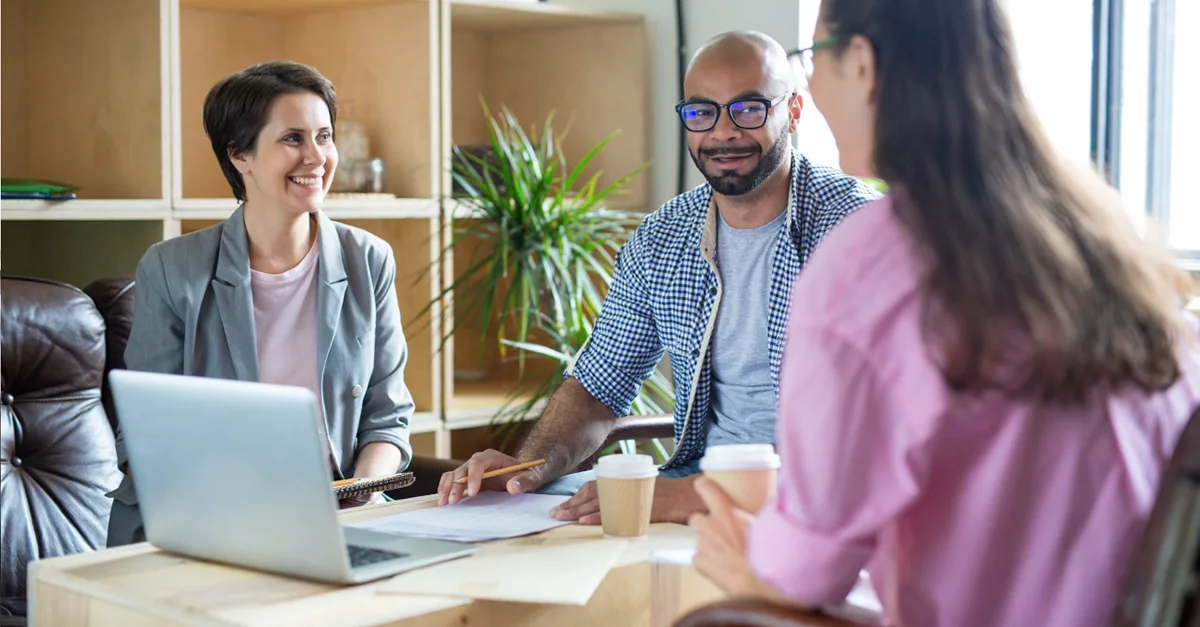 Three individuals sitting around a table in a modern office environment, engaged in a discussion with a laptop, papers, and drinks on the table.