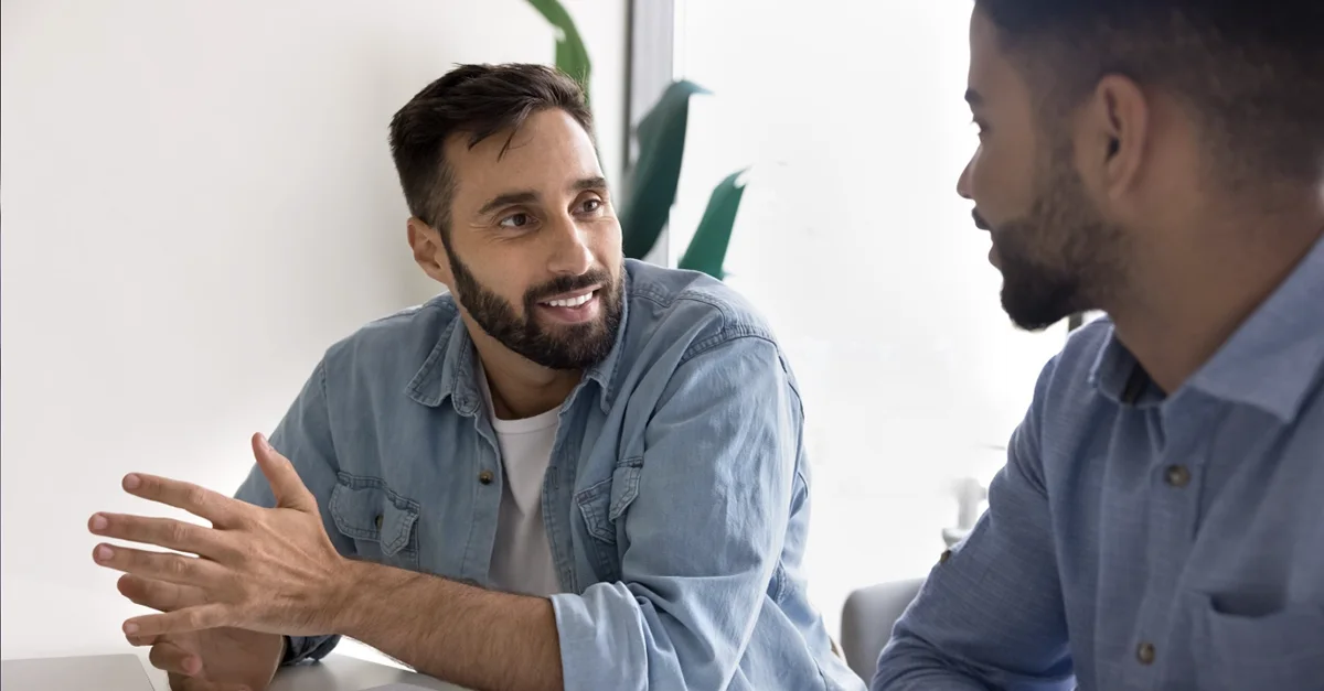 Two individuals seated at a desk having a discussion, with one person gesturing while a notebook, documents, and electronic devices are spread out on the table.